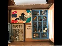 Box containing various colorful fan-shaped plastic earrings along with multiple pairs of gold-tone hoop and clip-on earrings, and blue earring organizer tray with multiple compartments.
