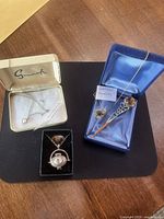 Four costume jewelry pieces displayed on a black background: pearl pendant in box, a silver and amber stone pin in box, a souvenir necklace in a black box, and a gold-filled hematite pendant on gold chain in a blue box.