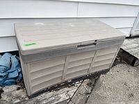 Large plastic outdoor deck box, light gray color, rectangular with slatted panel sides, placed outside on wooden pallets. Shows overall exterior condition.