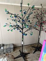 Full view of electric decorative tree with visible blue flowers and black metal branches, mounted on tripod base, standing against beige wall.