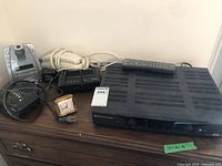 View of entire lot including black receiver/amplifier unit with remote, vintage travel alarm clock, telephone base, and other accessories on wooden surface