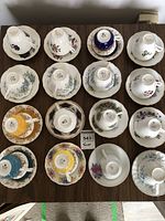 Overview shot showing all 16 teacups placed upside down on their matching saucers arranged in four rows on a table. Varied floral and ornate mid-20th century designs with multiple colors and gold trimming. Each teacup has the Royal Albert maker's mark visible on the bottom.