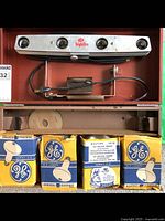 Silver EM Highlite light bar mounted horizontally in red box with attached cord and socket. Four boxes of GE bulbs and bulb accessories underneath.