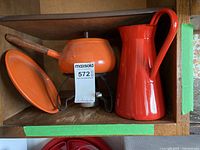 Three vintage kitchen items displayed in a wooden shelf: orange fondue pot with wooden handle, red enamel pitcher, and metal stand with burner and orange enamel plate.