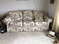 Full front view of floral upholstered sofa with two matching floral cushions on light carpeted floor with nearby small objects.