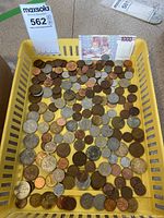 Basket with numerous mixed foreign coins, varying in size and color, with a 1000 lire banknote placed at the back inside the basket.