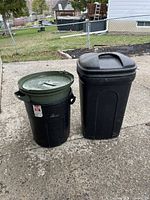 Photo showing both garbage cans side by side on concrete ground near a chainlink fence and yard.