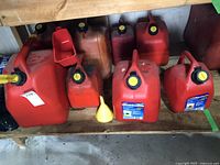 Red and orange plastic gas cans and two plastic funnels on a shelf. Various sizes are shown with black caps and yellow markings on some caps.