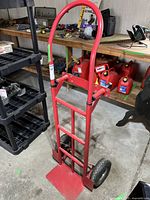 Front and side view of red hand cart showing metal frame, curved handle, and base plate.