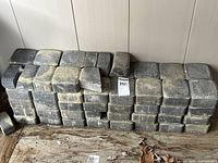 Pile of grey concrete interlocking bricks stacked against a wall with some yellow moss and dirt, showing weathered surfaces.