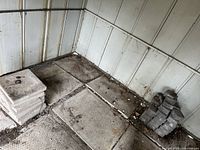 Concrete square slabs and stacked interlocking bricks on ground inside a metal shed, showing condition and size.