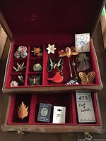 Photo showing the wooden jewelry box open to display the red velvet interior with assortment of costume brooches arranged in compartments including floral, butterfly, and bird shapes.