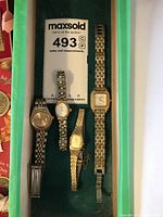 Overhead photo of four ladies wrist watches in different metal bands and face styles, laid out on green surface next to auction number card