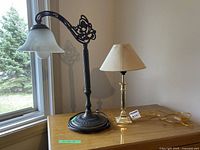 Two table lamps on a wooden surface near window and wall; one brass-finished with beige shade, one ornate dark metal with frosted glass shade.