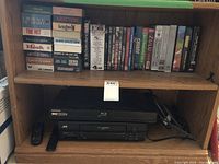Wooden shelf with assorted VHS and DVD movies on top shelf, Samsung Blu-ray DVD player and JVC VHS player with remote controls on lower shelves