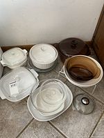 Photo of multiple white opaque Corning Ware casserole dishes with glass lids, a stack of clear glass bowls, a brown glass Pyrex bowl, and a separate glass lid on tile floor.
