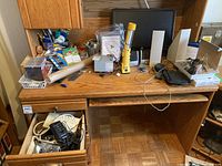 Overall view of wooden office desk covered with assorted office electronics, including monitor, speakers, cables, flashlight, computer mouse, and miscellaneous small items.