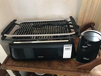 Philips HD6371 smokeless indoor electric grill with metal grilling surface, power cord visible. Black body with silver grill handles, placed on wooden chair.