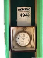 Top-down photo showing silver-tone Waltham pocket watch with white dial, black numerals, blue steel hands, sub-second dial, and emblem below 12 o'clock, set in a small box with cream padding.