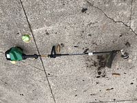 Side view of green and black Poulan FeatherLite SST25C gas trimmer lying on concrete showing entire length including handle and cutting head.