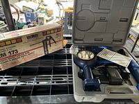 Photo showing the blue 7.2V power tool set flashlight and charger inside a grey plastic carrying case next to the boxed Lubrimatic PM-73 pneumatic grease gun.