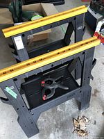 Two black folding saw horses with yellow top rails, folded open and standing on a concrete floor with a clamp on the shelf.