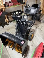 Full front-angle view of snow blower in garage, showing black/gray body and yellow wheels