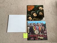 Three Beatles vinyl records displayed on carpet. Top right cover is 'Let It Be', bottom right is 'Sgt. Pepper's Lonely Hearts Club Band', left is the plain white cover of the 'White Album'