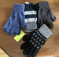 Photo shows four pairs of knit gloves on a wooden surface: blue speckled, navy white striped, solid grey, and black with geometric pattern.