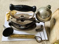 Antique self heating gas powered iron with black handle, embodied text 'Comfort Iron' and 'Self Heating', spherical metal fuel container attached on side, brass funnel and small tool nearby all on carpet.