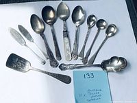 Overview photo of all 11 spoons and butter spreaders laid out on white background with handwritten note '133 Antique 11x silver plate spoons'