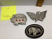 Three belt buckles shown front side: pig-shaped metal buckle with red hearts and text, wing-shaped rhinestone star buckle, oval leather horse buckle.
