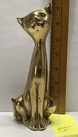Front view of the brass cat figurine placed against a ruler for scale, showing approx. 6 inch height and stylized facial features.