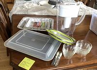 Overall lot showing cake pan with lid, foil pans, water pitcher, glass bowls, butter spreaders, and collapsible strainer