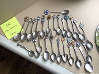 Collection of 35 collector spoons laid out on a white surface with a yellow note showing quantity 35 and mention of couple missing pieces.
