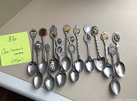 Complete overview of 14 Canadian souvenir collector spoons arranged in a row, showing various handles with colorful emblems, one spoon missing part of the handle decoration.
