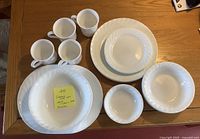 Front view of the dishes laid out including bowls, plates, and four mugs showing the swirl pattern on the edges of the white dishes.