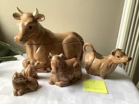 Front view of the ceramic cow tableware set showing all pieces assembled and displayed on a tablecloth, including large covered dish, creamer, sugar bowl, salt and pepper shakers.