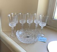 Six crystal wine glasses arranged in a row with thick bottoms and cut patterns, plus a small round crystal dish featuring detailed cut designs and scalloped edge, all displayed on a white surface near a window.