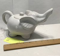 Elephant-shaped white ceramic tea pot shown from an angled side view with a ruler for scale, about 8 inches long.