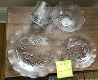 Pressed glass dishes including large dinner plate, smaller salad plate, dessert bowl, and a clear glass mug with floral pressed design, all arranged on a wooden surface.