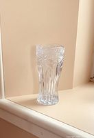 Full view of the tall clear pressed glass vase with frosted rim and floral pattern, placed against a plain wall on a surface.