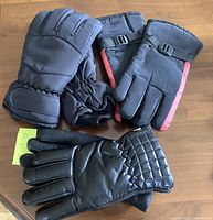Three pairs of men's gloves displayed on a wooden surface: two insulated winter gloves including one pair with red stripe and adjustable strap, and one pair of black faux leather gloves with quilted wrist detail.