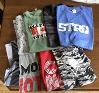 Eight men's T-shirts in size XL, assorted colors and graphics, laid flat on wooden surface.
