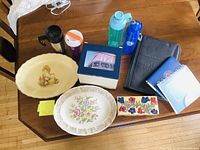 Overview of lot on wooden table including antique platters, wooden boxes, water bottles, travel mug, zippered portfolio, notebooks and papers