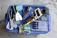 Photo showing variety of hand tools including hammers, scrapers, pliers, hacksaw, wire, and hardware items on blue bin lid