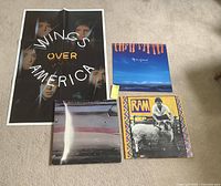 Photo showing two vinyl records 'Ram' and 'Off the Ground', Wings Over America poster and card print on floor.