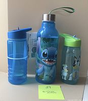 Photo of three refillable water bottles: a blue bottle with a flip spout lid, a blue metal-capped bottle with Stitch graphic and wrist strap, and a clear bottle with a green lid and cartoon characters.