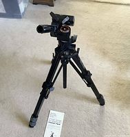Photo of the fully extended black Slik tripod standing on carpeted floor with operating instructions in front.