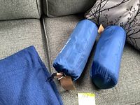 Two navy blue plush blankets rolled and stored in blue carry sacks placed on a gray couch with a floral pillow in the background.